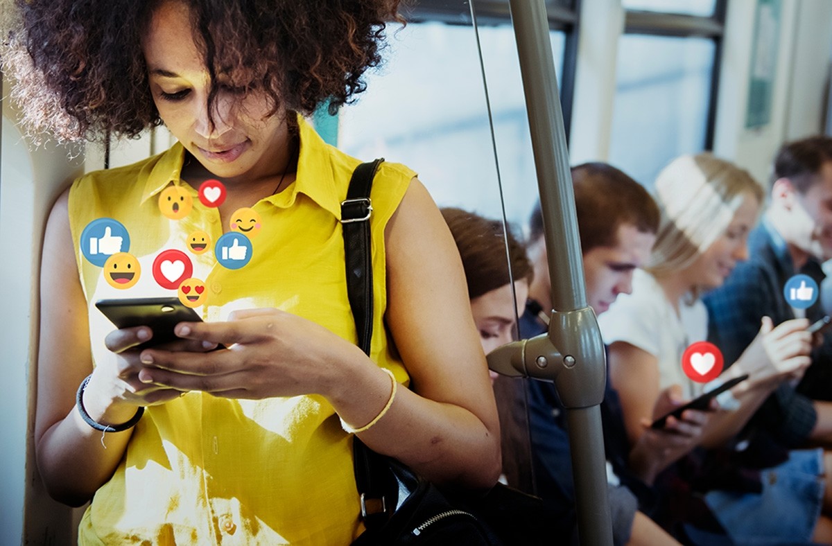 Woman in yellow blouse standing on public transportation using social media on her smartphone with 4 other people in background using social media