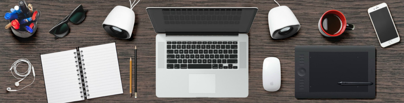 Desk filled by notepad, writing utensils, headphones, sunglasses, pencil cup, laptop, mouse, speakers, coffee, phone, and tablet