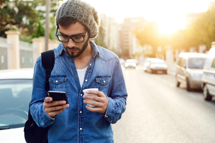 Man standing in front of car, wearing a beanie and backpack, coffee in hand, using mobile device