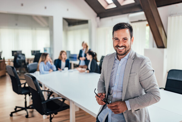 Man in business casual attire smiling and standing in front of long white meeting table with digital marketing business associates sitting in background